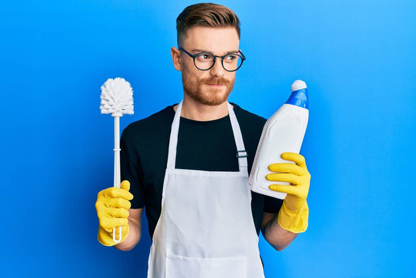 Junger rothaariger Mann macht Badezimmer-Hausarbeit mit Klobürste lächelnd zur Seite schauen und wegstarren denken.  - Foto, Bild