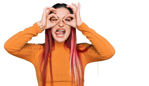 Young caucasian woman wearing casual clothes doing ok gesture like binoculars sticking tongue out, eyes looking through fingers. crazy expression.  - Photo, Image