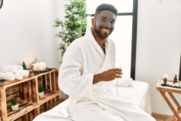 Young african american man smiling happy with facial mask drinking champagne at beauty center. - Fotó, kép