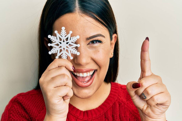 Bella donna bruna che tiene il fiocco di neve sopra l'occhio sorridente con un'idea o una domanda che punta il dito con la faccia felice, numero uno  - Foto, immagini