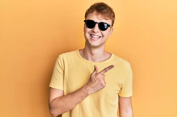 Junger kaukasischer Mann mit stylischer Sonnenbrille lächelt fröhlich und zeigt mit der Hand und dem Finger zur Seite  - Foto, Bild