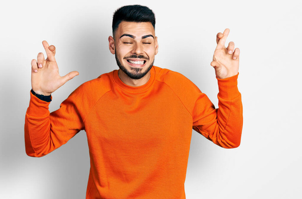 Молодой латиноамериканец с бородой в обычном оранжевом свитере, жестикулирующий пальцем, улыбающийся с надеждой и закрытыми глазами. удача и суеверие.  - Фото, изображение