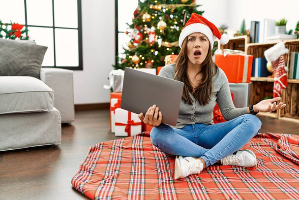 Jeune femme latine utilisant un ordinateur portable assis près de l'arbre de Noël dans le visage de choc, l'air sceptique et sarcastique, surpris avec la bouche ouverte  - Photo, image