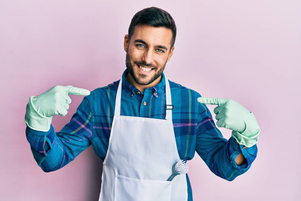 Young hispanic man wearing apron looking confident with smile on face, pointing oneself with fingers proud and happy.  - Фото, зображення