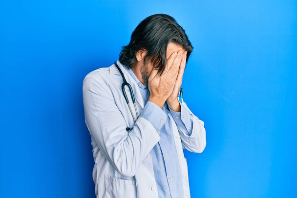 Pohledný muž středního věku v doktorské uniformě a stetoskopu se smutným výrazem zakrývajícím obličej rukama při pláči. deprese koncept.  - Fotografie, Obrázek