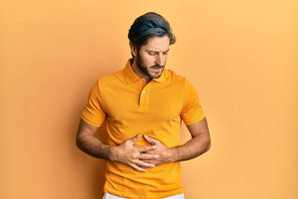 Hombre hispano joven con camiseta amarilla casual con la mano en el estómago porque la indigestión, la enfermedad dolorosa sensación de malestar. concepto ache.  - Foto, imagen
