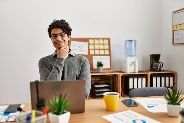 Junger hispanischer Mann im Business-Stil, der am Schreibtisch im Büro sitzt und selbstbewusst in die Kamera lächelt, mit verschränkten Armen und erhobener Hand am Kinn. Positives Denken.  - Foto, Bild