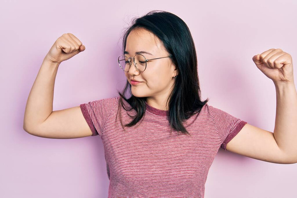 Joven chica china con ropa casual y gafas que muestran los músculos de los brazos sonriendo orgulloso. concepto de fitness.  - Foto, Imagen