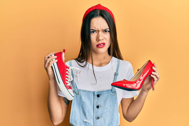 Молода іспаномовна дівчина вибирає туфлі на підборах і кросівки скептично і нервово, розчаровуючись через проблеми. негативна людина
.  - Фото, зображення