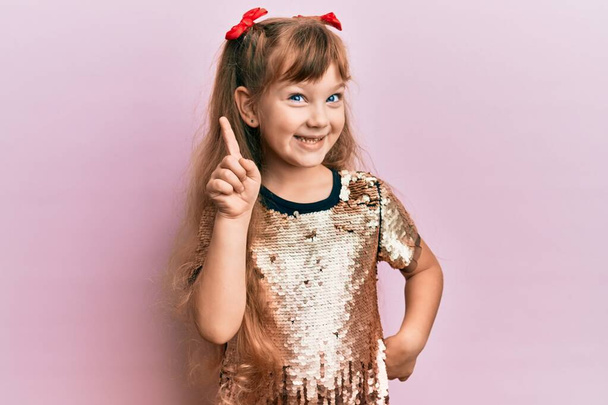 Petite fille caucasienne enfant portant robe de paillettes festives montrant et pointant vers le haut avec le doigt numéro un tout en souriant confiant et heureux.  - Photo, image
