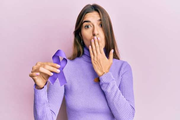 Nuori latino nainen tilalla violetti nauha tietoisuutta kattaa suun käsin, järkyttynyt ja pelkää virhe. yllättynyt ilme  - Valokuva, kuva