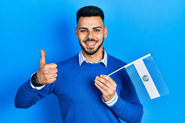 Молодой латиноамериканец с бородой с флагом Сальвадора улыбается счастливым и позитивным, большой палец вверх делает отлично и знак одобрения  - Фото, изображение