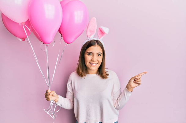 Młoda piękna kobieta nosi słodkie uszy króliczka wielkanocnego i trzyma balony uśmiechając szczęśliwy wskazując ręką i palcem na bok  - Zdjęcie, obraz