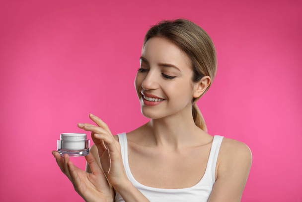 Молодая женщина держит банку крема для лица на розовом фоне - Фото, изображение