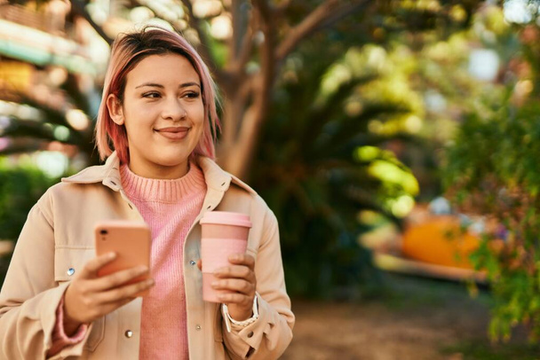 Νεαρή ισπανόφωνη κοπέλα που χρησιμοποιεί smartphone και πίνει καφέ στην πόλη. - Φωτογραφία, εικόνα