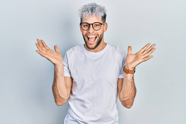 Jeune homme hispanique aux cheveux teints modernes portant un t-shirt blanc et des lunettes célébrant fou et étonné pour le succès avec les bras levés et les yeux ouverts hurlant excités. concept gagnant  - Photo, image