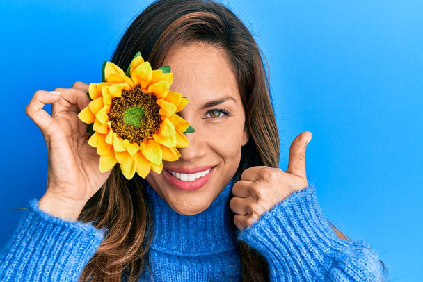 Mladá latinská žena drží slunečnici přes oko usměvavý šťastný a pozitivní, palec nahoru dělá vynikající a schvalovací znamení  - Fotografie, Obrázek