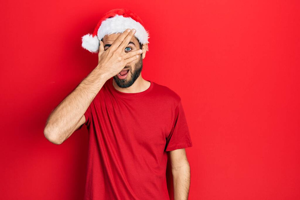 若いですヒスパニック男身に着けていますクリスマスの帽子の目ショックで顔と目をカバー手で,恥ずかしい表情で指を通して見る.  - 写真・画像