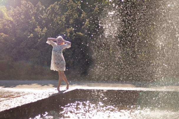 Attractive girl in white short dress standing near fountain at the summer hottest day. Girl with dress partly wet dancing. Beautiful blonde women near the fountain in a ballet position - Photo, Image