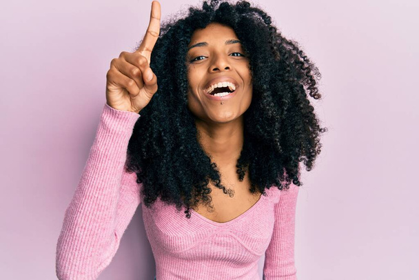 Mujer afroamericana con cabello afro que usa una camisa rosa casual apuntando con el dedo hacia arriba con una idea exitosa. exitado y feliz. número uno.  - Foto, imagen