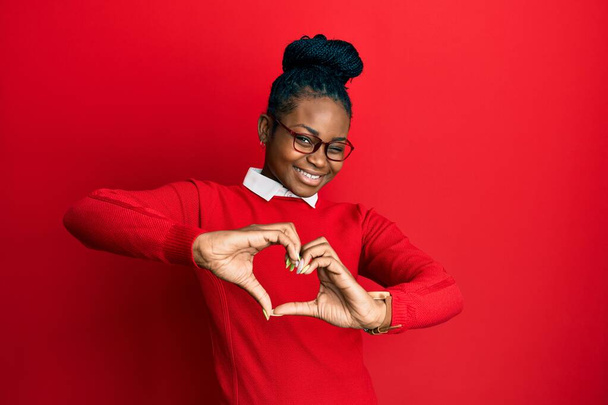 Fiatal afro-amerikai nő alkalmi ruhát és szemüveget visel, szerelmesen mosolyog, miközben szív szimbólum alakot ölt a kezével. romantikus koncepció.  - Fotó, kép