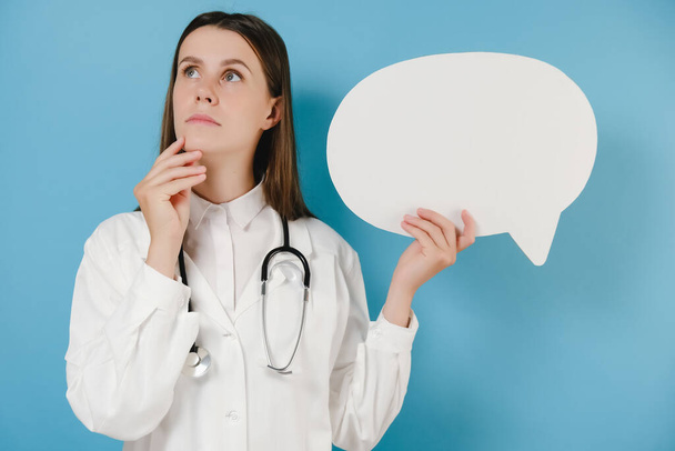 Pensive young doctor woman in white coat and stethoscope hold empty blank Say cloud speech bubble, isolated on blue background. Epidemic pandemic coronavirus 2019-ncov sars covid-19 flu virus concept - Fotografie, Obrázek