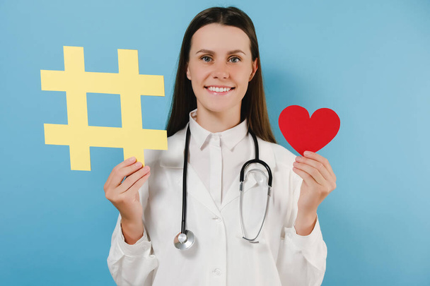 Portrait of friendly young woman doctor with toothy smile have good mood holding large big yellow hashtag sign and little red heart, wears coat, posing isolated over blue color background in studio - Фото, зображення