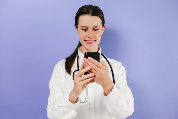 Portrait of happy young doctor lady hold smartphone look screen wear stethoscope and white uniform, online prescription network website innovation, posing isolated over purple studio background wall - Фото, зображення