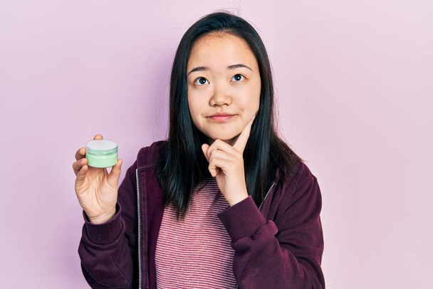 Молодая китаянка держит косметический увлажняющий крем для лица серьезное лицо думая о вопросе с рукой на подбородке, вдумчивый о запутанной идее  - Фото, изображение