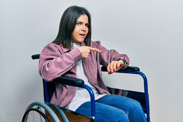 Young brunette woman sitting on wheelchair in hurry pointing to watch time, impatience, upset and angry for deadline delay  - Foto, Imagen