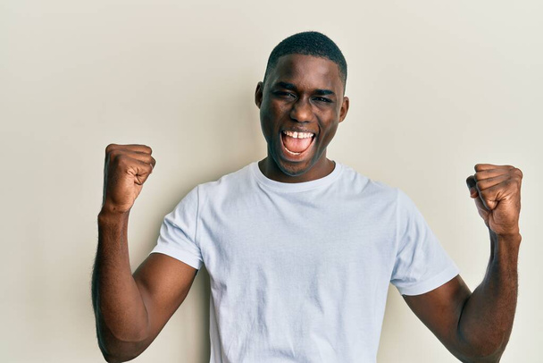 Jonge Afro-Amerikaanse man draagt casual wit t-shirt schreeuwen trots, vieren overwinning en succes zeer enthousiast met opgeheven armen  - Foto, afbeelding