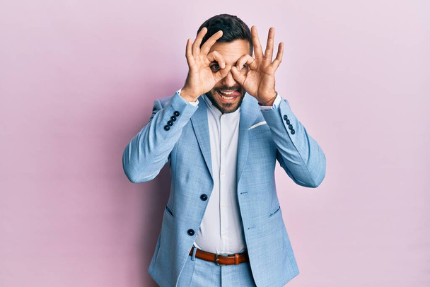 Young hispanic businessman wearing business jacket doing ok gesture like binoculars sticking tongue out, eyes looking through fingers. crazy expression.  - Foto, Bild