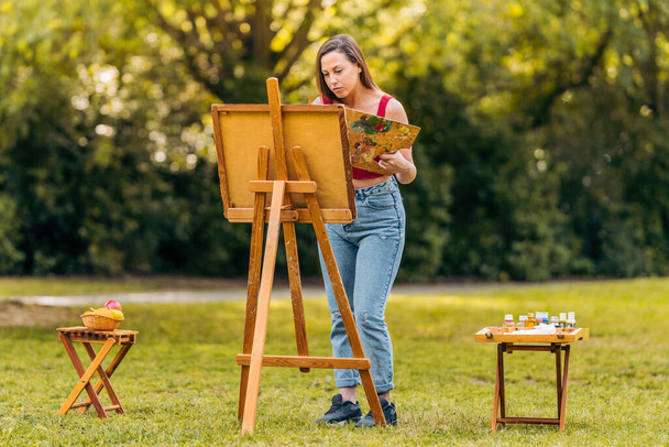 Mujer pintando sobre lienzo en un parque con dos mesas auxiliares con herramientas - Foto, Imagen