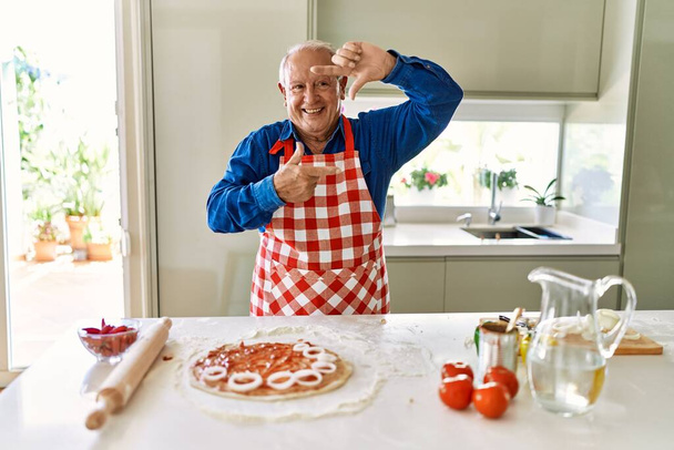 Senior man with grey hair cooking pizza at home kitchen smiling making frame with hands and fingers with happy face. creativity and photography concept.  - Fotó, kép