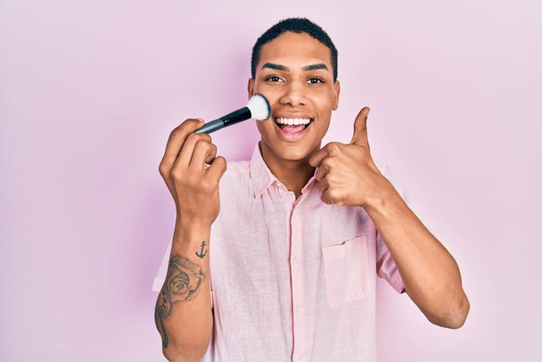 Young african american guy holding makeup brush smiling happy and positive, thumb up doing excellent and approval sign  - Foto, imagen
