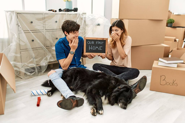 Jeune couple caucasien avec chien tenant notre premier tableau noir à la maison dans une nouvelle maison rire et rire embarrassé couvrant la bouche avec les mains, commérages et concept de scandale  - Photo, image