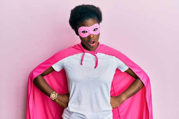 Молодая африканская американская девушка в костюме супергероя и медицинской маске в шокирующем лице, выглядящая скептически и саркастично, удивленная открытым ртом  - Фото, изображение