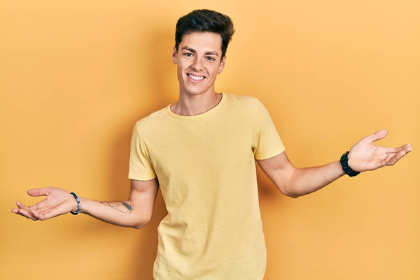 Giovane uomo ispanico indossa casual t shirt gialla sorridente mostrando entrambe le mani palme aperte, presentando e pubblicizzando confronto ed equilibrio  - Foto, immagini