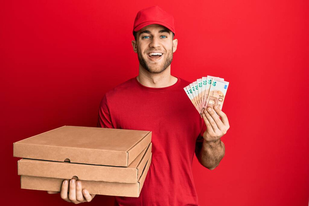 Young caucasian man holding take away food and 10 euros banknotes smiling and laughing hard out loud because funny crazy joke.  - Fotografie, Obrázek