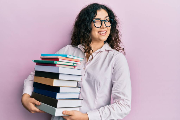 Young brunette woman with curly hair holding a pile of books winking looking at the camera with sexy expression, cheerful and happy face.  - Fotó, kép