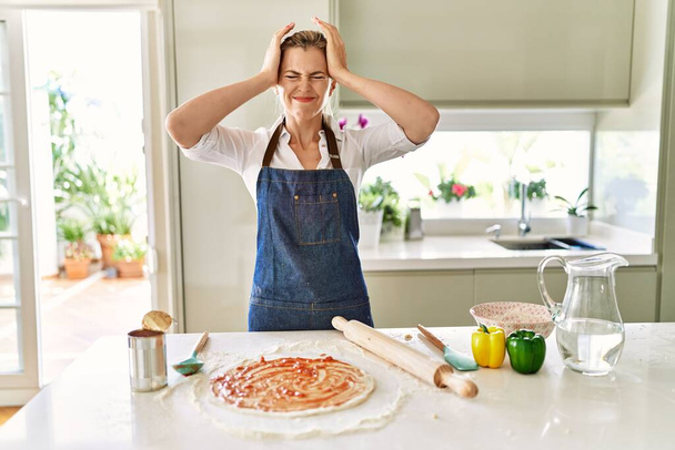 Schöne blonde Frau in Schürze Pizza kochen leiden unter Kopfschmerzen verzweifelt und gestresst, weil Schmerzen und Migräne. Hände auf den Kopf.  - Foto, Bild