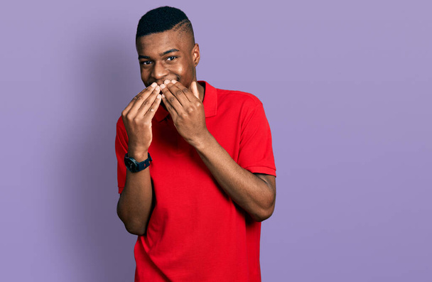Jonge Afro-Amerikaanse man draagt casual rood t-shirt lachen en beschaamd giechelen bedekken mond met handen, roddel en schandaal concept  - Foto, afbeelding