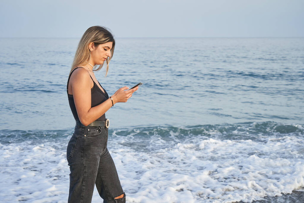 Nuori nainen kävelee rannalla älypuhelimen kanssa - Valokuva, kuva