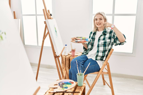 Sanat stüdyosunda resim yapan genç sanatçı kadın neşeli bir şekilde gülümseyip parmaklarını ve ağzını işaret ediyor. diş sağlığı kavramı.  - Fotoğraf, Görsel