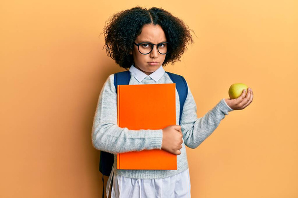 Młoda dziewczynka z afro włosy noszące torbę szkolną trzymając książki i zielone jabłko sceptyczny i nerwowy, marszczenie brwi zdenerwowany z powodu problemu. osoba negatywna.  - Zdjęcie, obraz