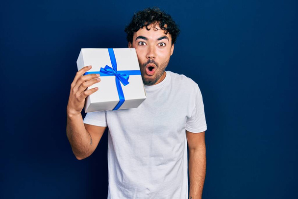 Joven hombre hispano sosteniendo regalo asustado y sorprendido con la boca abierta para sorpresa, cara de incredulidad  - Foto, imagen