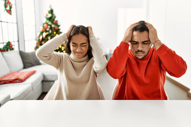 Noel ağacının yanında masada oturan genç İspanyol çift acı ve migren yüzünden çaresiz ve stresli bir baş ağrısından muzdarip. Eller başının üstünde..  - Fotoğraf, Görsel