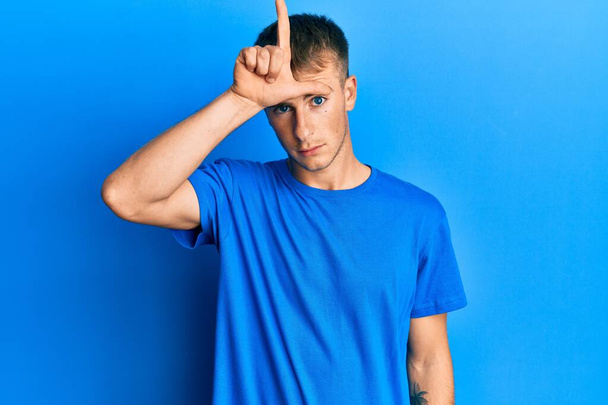 junger kaukasischer Mann in lässigem blauem T-Shirt, der sich über Menschen mit Fingern auf der Stirn lustig macht, indem er die Geste des Verlierers verspottet und beleidigt.  - Foto, Bild