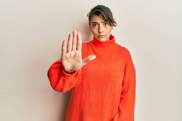 Jonge Spaanse vrouw draagt casual winter trui doen stoppen met zingen met palm van de hand. waarschuwingsuitdrukking met negatief en ernstig gebaar op het gezicht.  - Foto, afbeelding