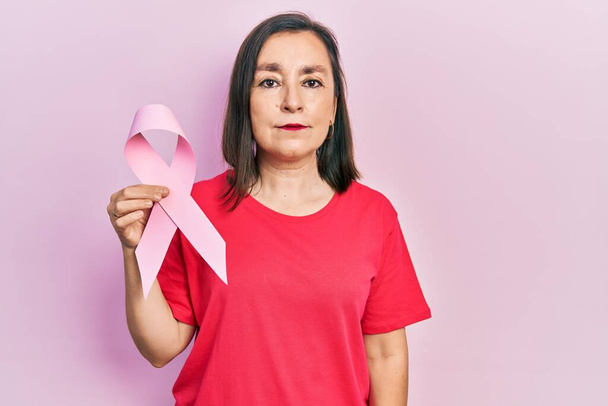 Middle age hispanic woman holding pink cancer ribbon thinking attitude and sober expression looking self confident  - Foto, Imagem